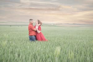 Portrait romantique d'un couple en séance d'engagement pris par SL-Photographie, avec un homme en chemise rouge à pois tenant tendrement une femme en robe rouge éclatante, tous deux souriants et se regardant avec affection, entourés par un champ de blé verdoyant avec un ciel nuageux au crépuscule en arrière-plan, photographié à Courtisols.