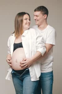Couple souriant de futurs parents, en tenues décontractées blanches, moment tendre partagé autour du ventre enceinte, par SL-Photographie.