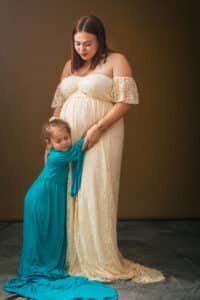 Une future maman rayonnante portant une robe de grossesse en dentelle ivoire, accompagnée de sa jeune fille câlinant affectueusement son ventre rond, immortalisées par SL-Photographie dans leur studio à Châlons-en-Champagne.