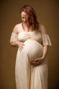 Portrait intimiste et chaleureux d'une femme enceinte aux cheveux auburn, étreignant son ventre enveloppé dans une élégante robe ivoire, capturé par SL-Photographie à Châlons-en-Champagne.