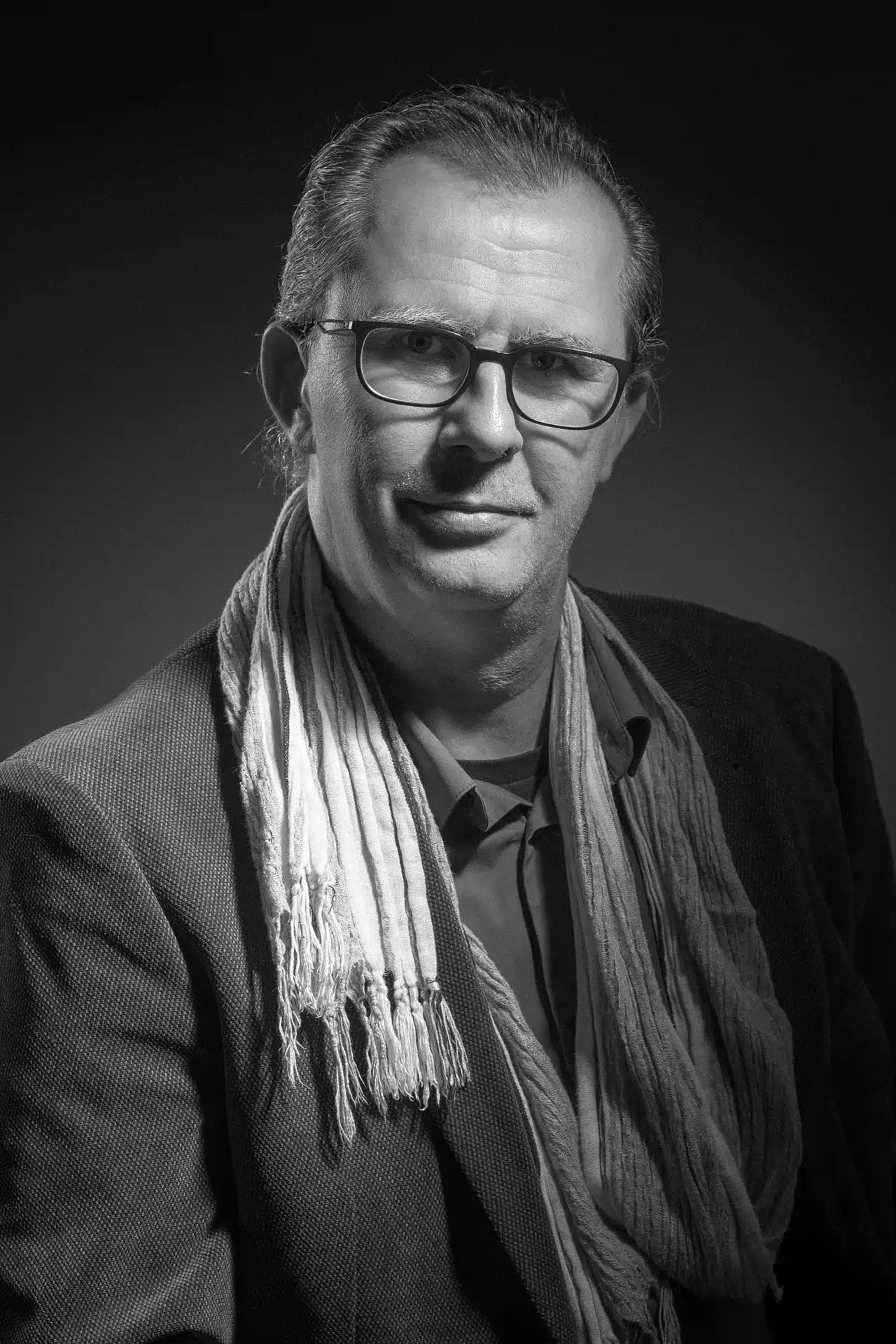SL-Photographie-Sebastien-Loppin-portrait-portrait-autoportrait Autoportrait en noir et blanc d'un homme avec des lunettes, souriant légèrement et portant une veste sombre et une écharpe claire, réalisé par le photographe dans son studio à Courtisols, exprimant une personne professionnelle et accessible.
